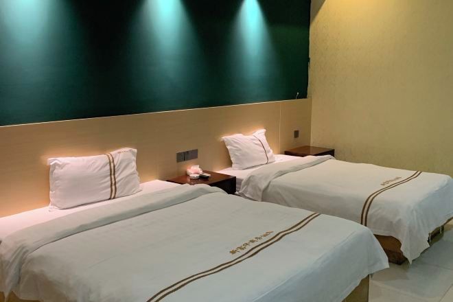 Una habitación de hotel con dos camas, cada una con cubrecama y almohadas blancas, una mesita de noche con lámpara y un televisor de gran tamaño montado en la pared.