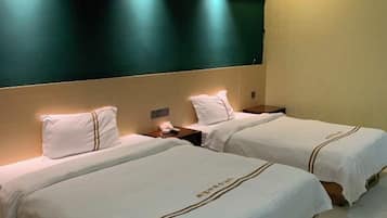 Una habitación de hotel con dos camas, cada una con cubrecama y almohadas blancas, una mesita de noche con lámpara y un televisor de gran tamaño montado en la pared.