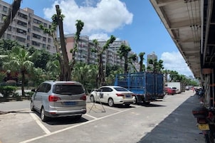 Un área de estacionamiento con vehículos, incluyendo un automóvil blanco y una camioneta azul, y un edificio de varios pisos al fondo.