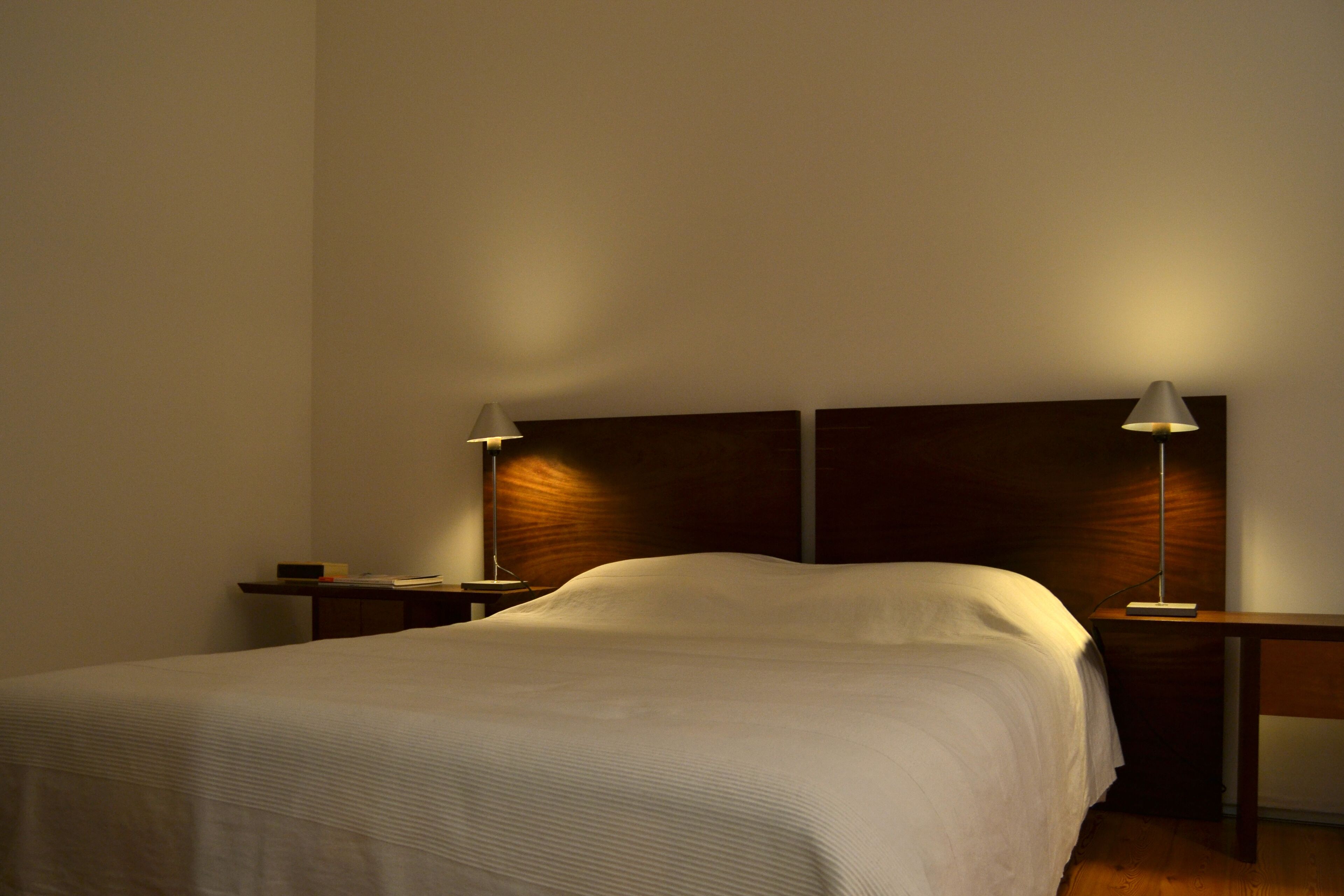 A neatly made bed with a wooden headboard, two bedside tables with lamps, and a plain wall.
