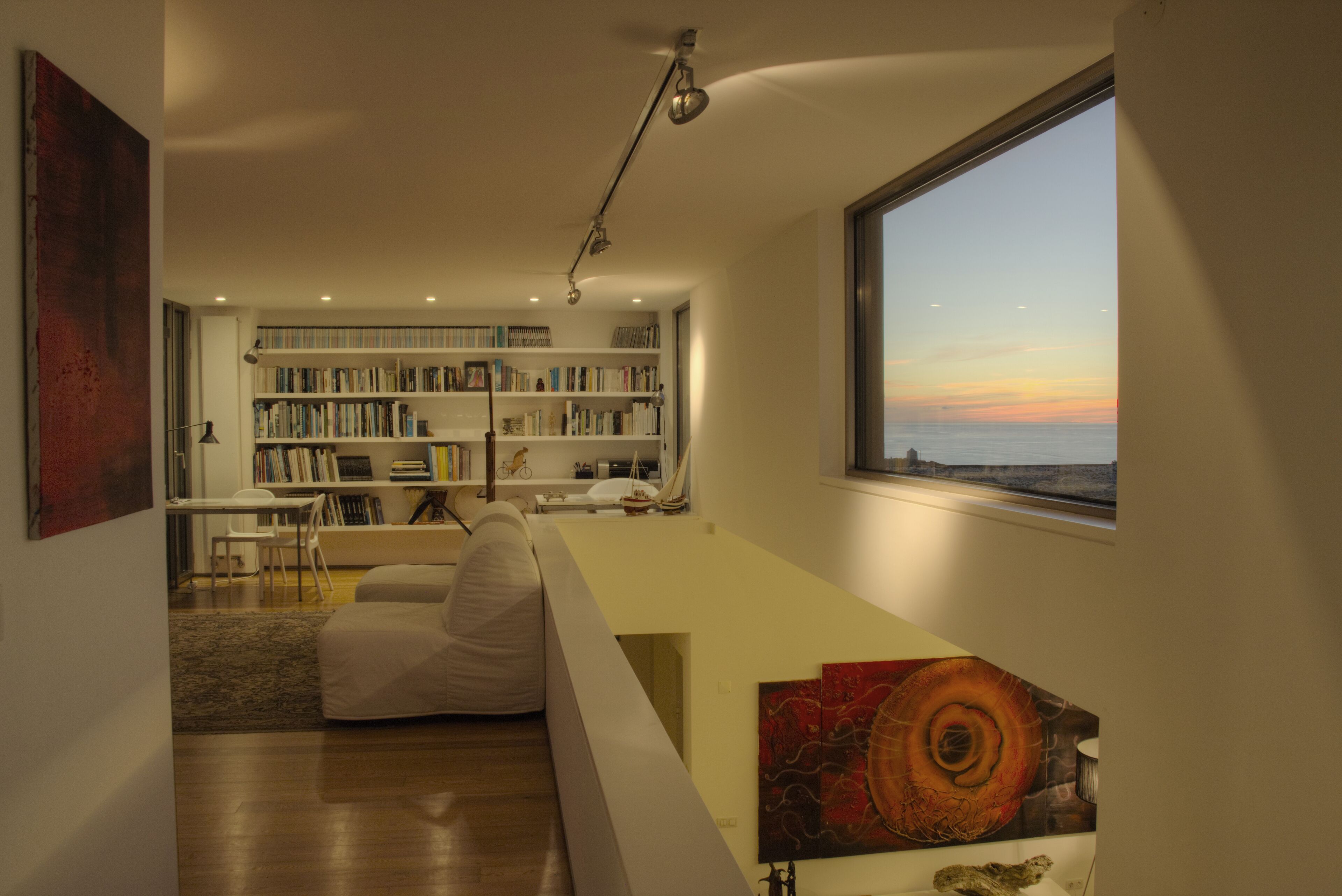 A modern living room with a large bookshelf, a white sofa, and a view of the sunset through a large window.