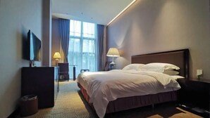 Une chambre d’hôtel avec un grand lit, un bureau avec une lampe, une télévision et une fenêtre avec des rideaux.