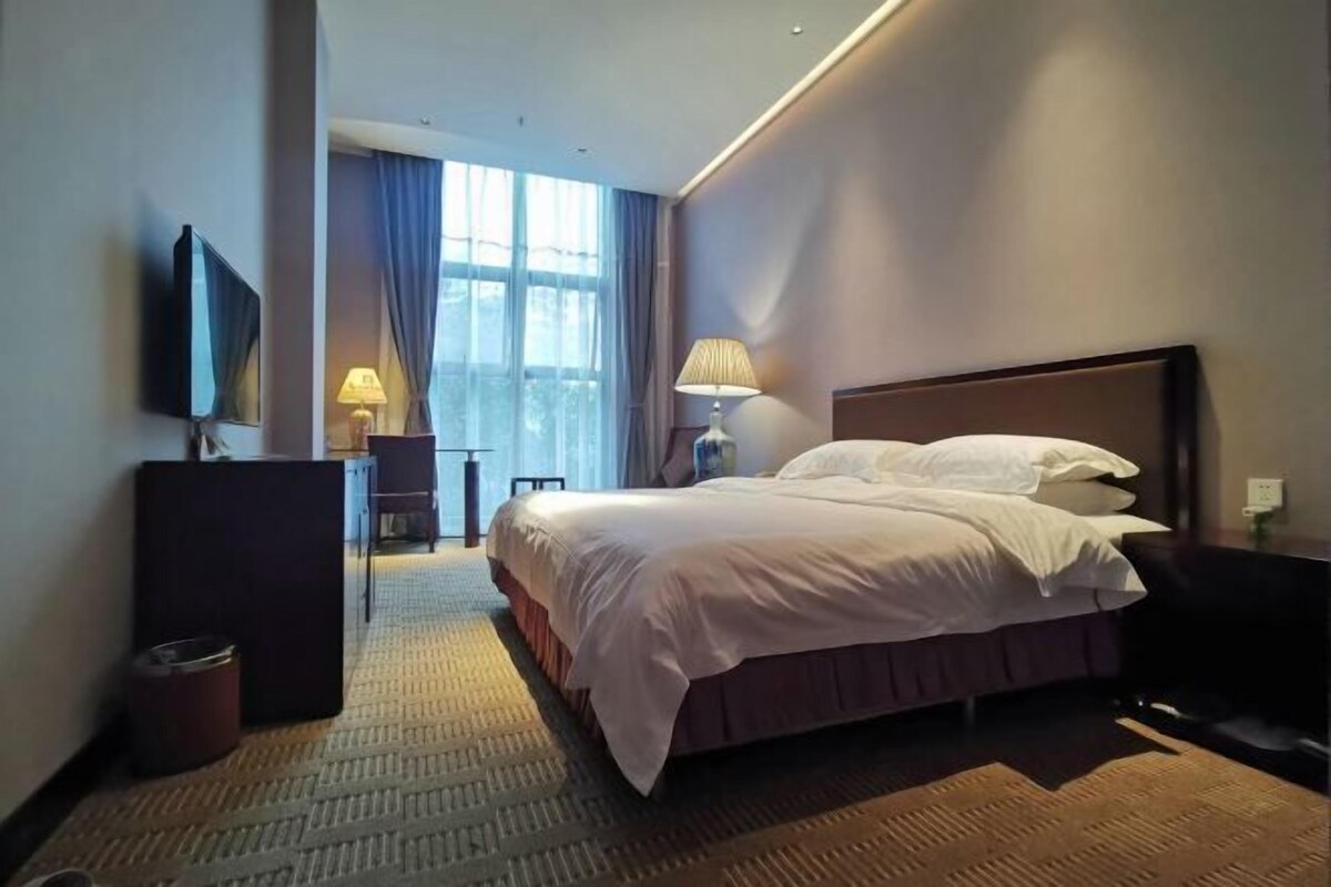 Una habitación de hotel con una cama grande, un escritorio con lámpara, un televisor y una ventana con cortinas.