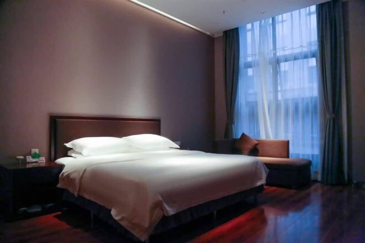 A hotel room with a large bed, a nightstand, a chair, and a window with sheer curtains.