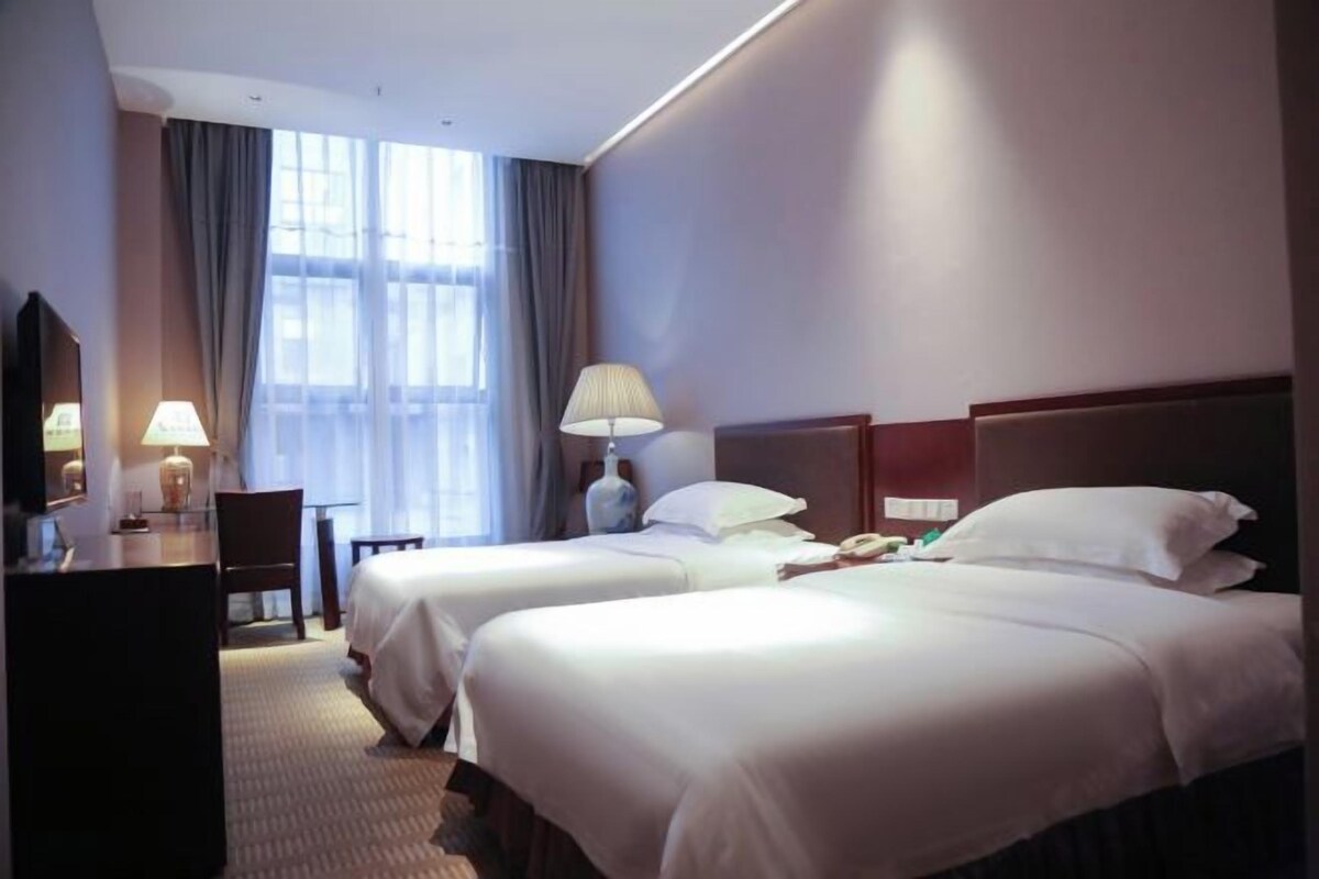 A hotel room with two beds, a desk, a chair, a lamp, and a large window with curtains.
