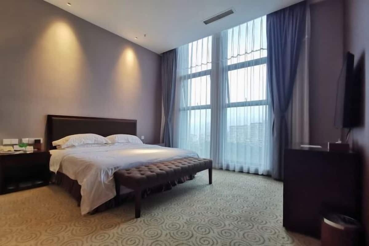 A hotel room with a large bed, a bench, a TV, and a window with curtains.