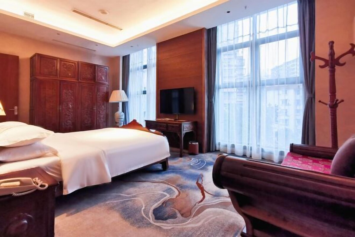 Una habitación de hotel con cama, escritorio, televisor y un amplio ventanal con cortinas.