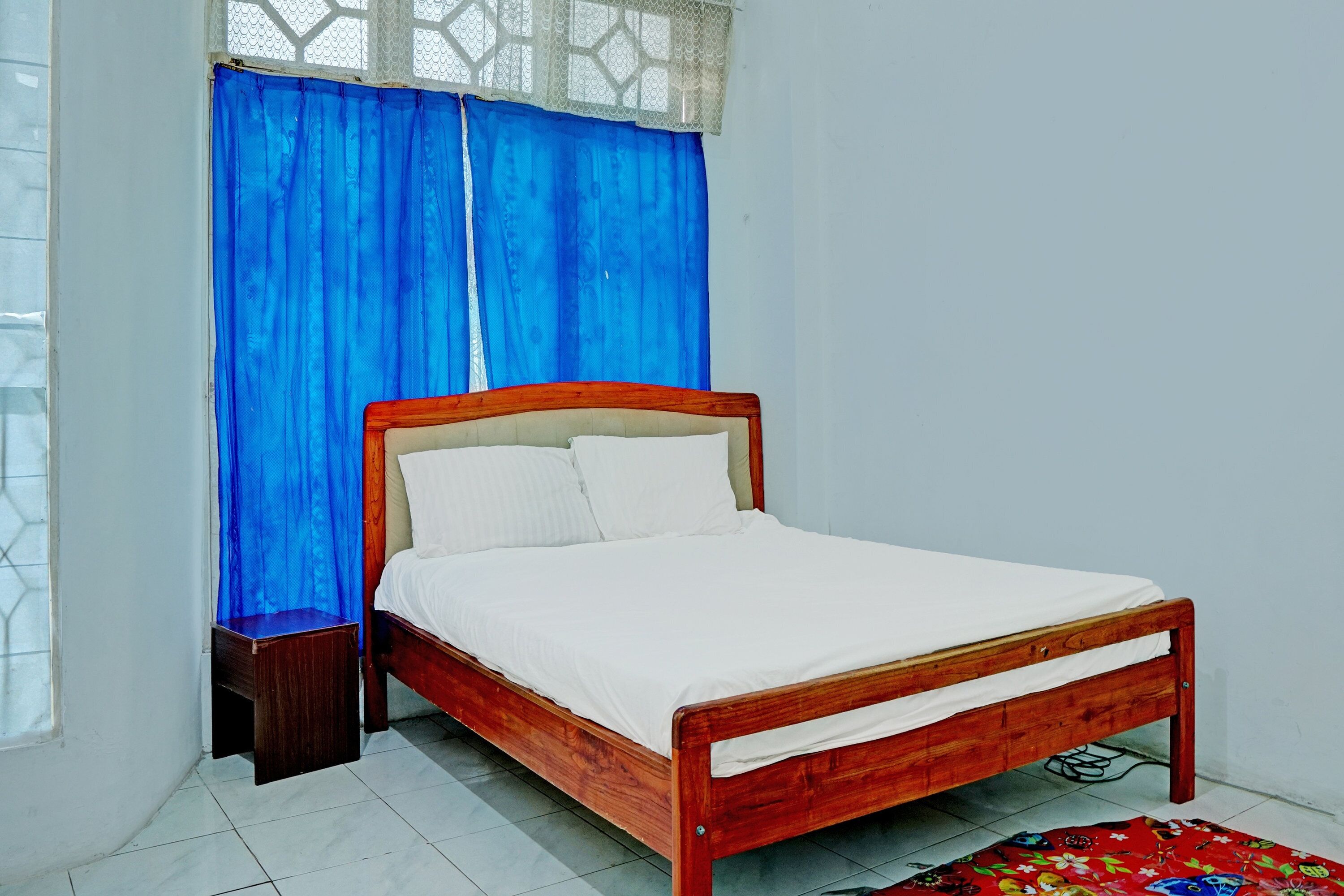A single bed with a wooden frame and white bedding in a room with blue curtains and a patterned rug.