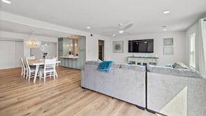 A modern living room with a grey sectional sofa, a dining table with chairs, a kitchen area with a TV, and a ceiling fan. - Relax in Beachside Comfort with Stunning Views (Galveston)