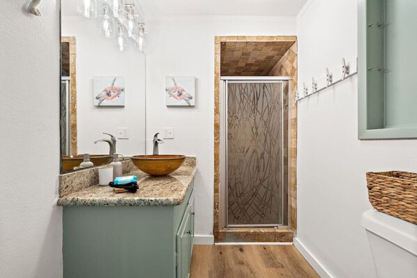 Une salle de bain dotée d’un comptoir en granit, d’un lavabo en forme de bol en bois, d’une douche à parois de verre et d’un miroir orné d’une œuvre d’art.