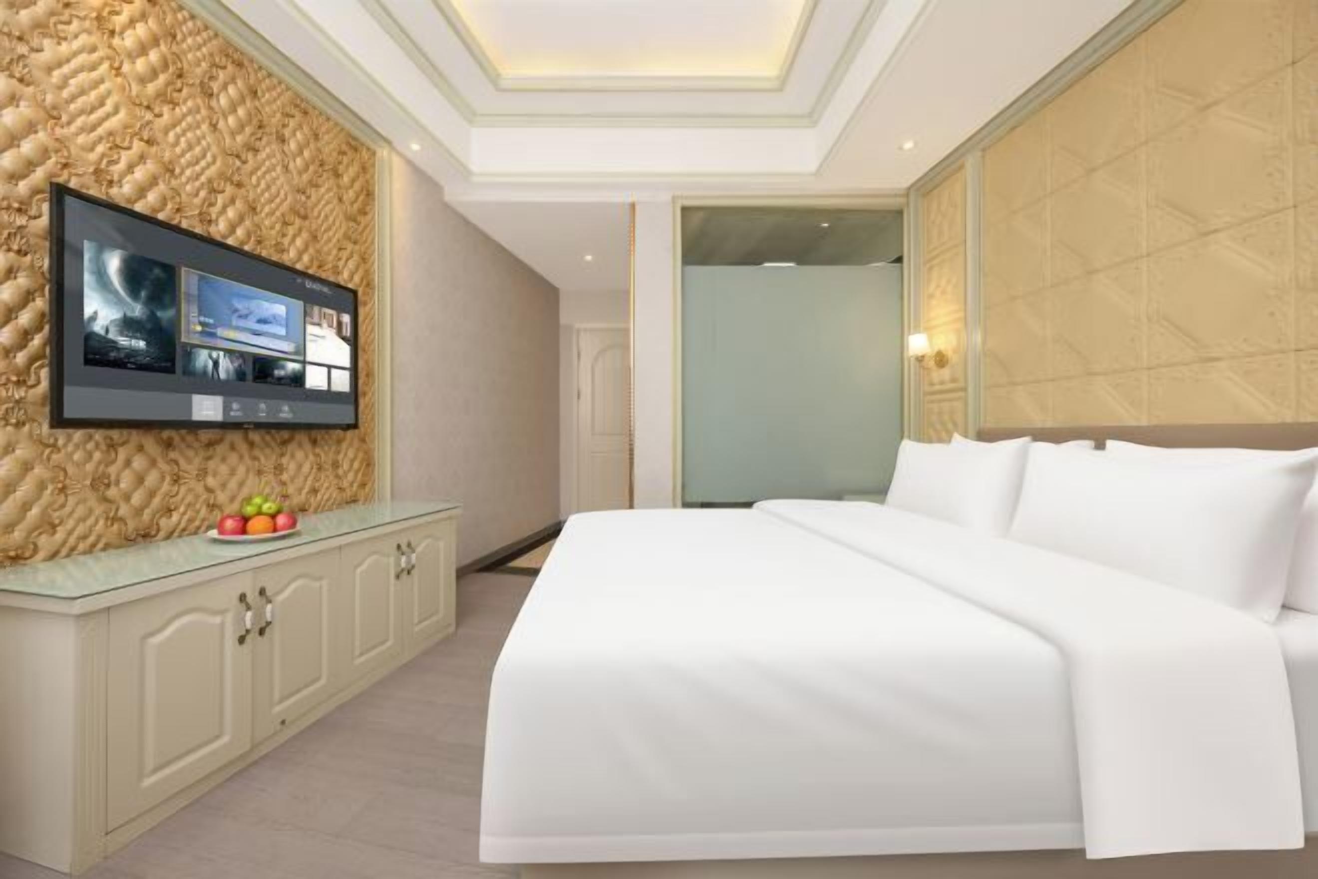 A modern hotel room with a large bed, a flat-screen TV mounted on a textured wall, and a glass partition separating the room from another space.