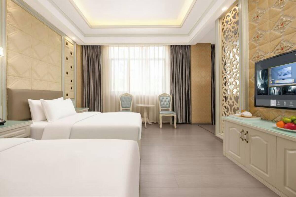 A hotel room with two beds, a TV, a small table with fruit, and a decorative wall panel.