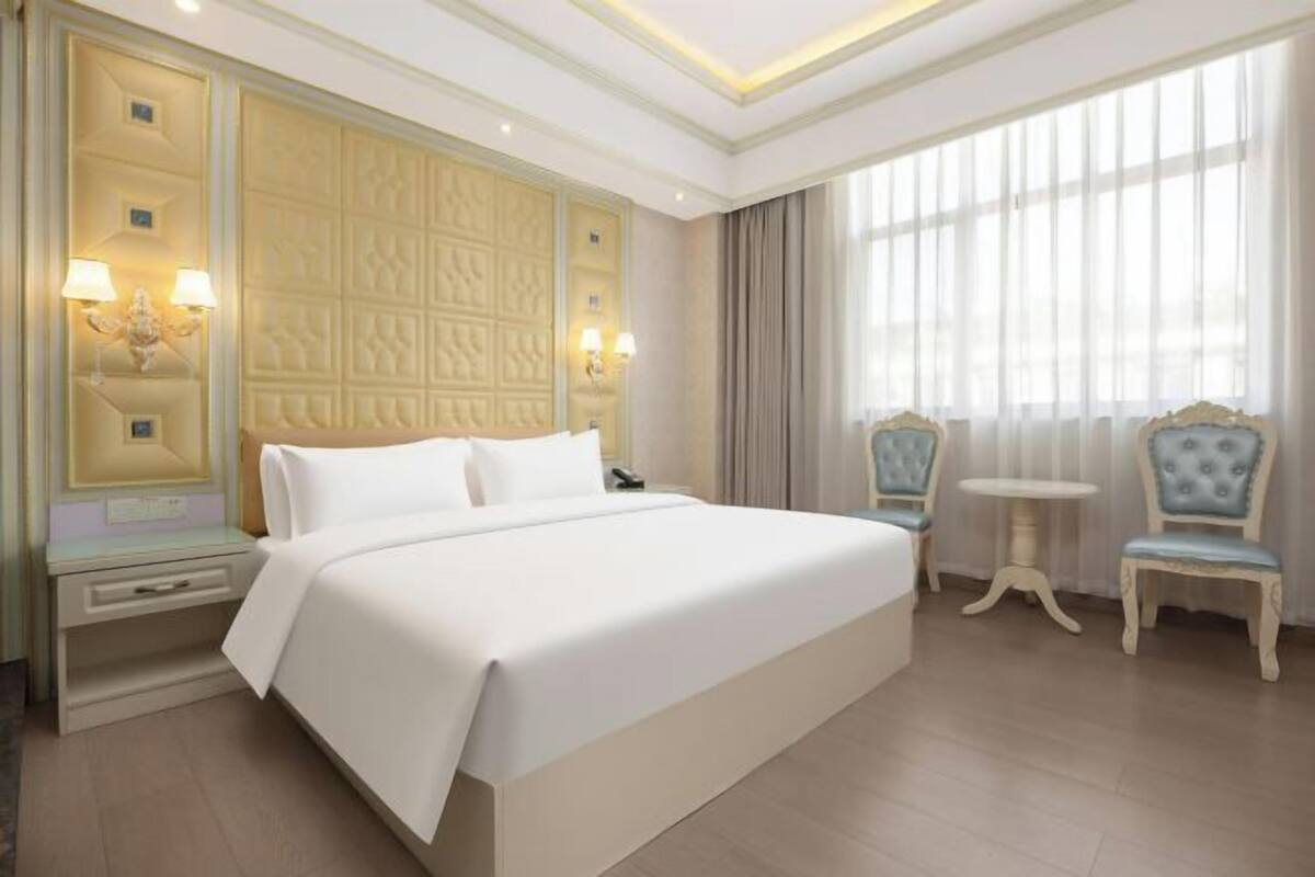 A hotel room with a large bed, bedside tables, a nightstand, two chairs, and a window with curtains.