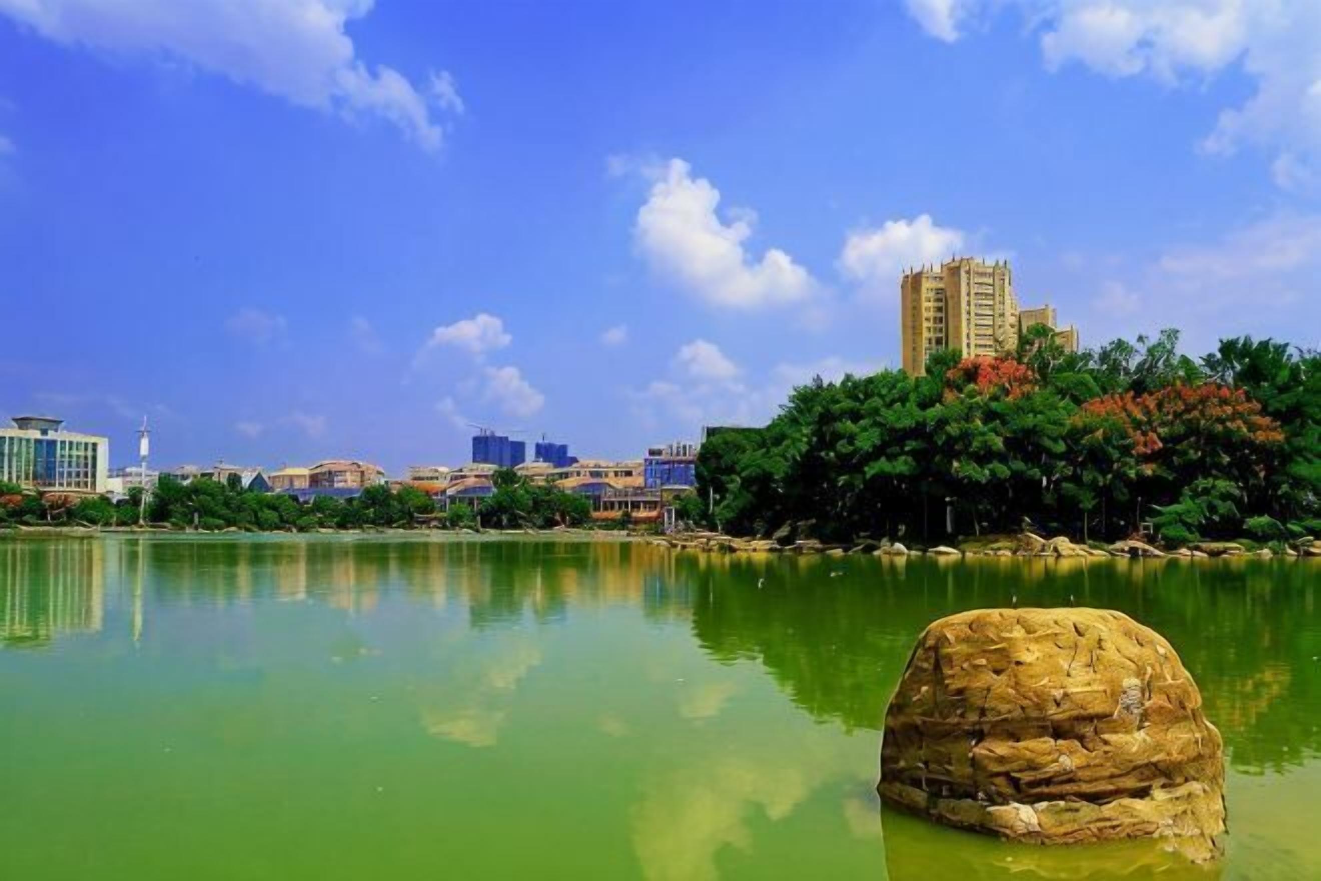 A calm lake with a large rock in the foreground, a building with a tower on the right, and a clear blue sky.