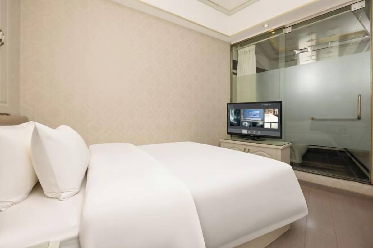 A hotel room with a bed, a television, a glass shower enclosure, and a white wall.