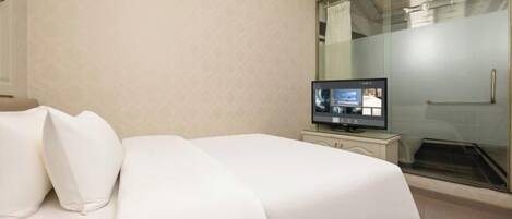 A hotel room with a bed, a television, a glass shower enclosure, and a white wall.