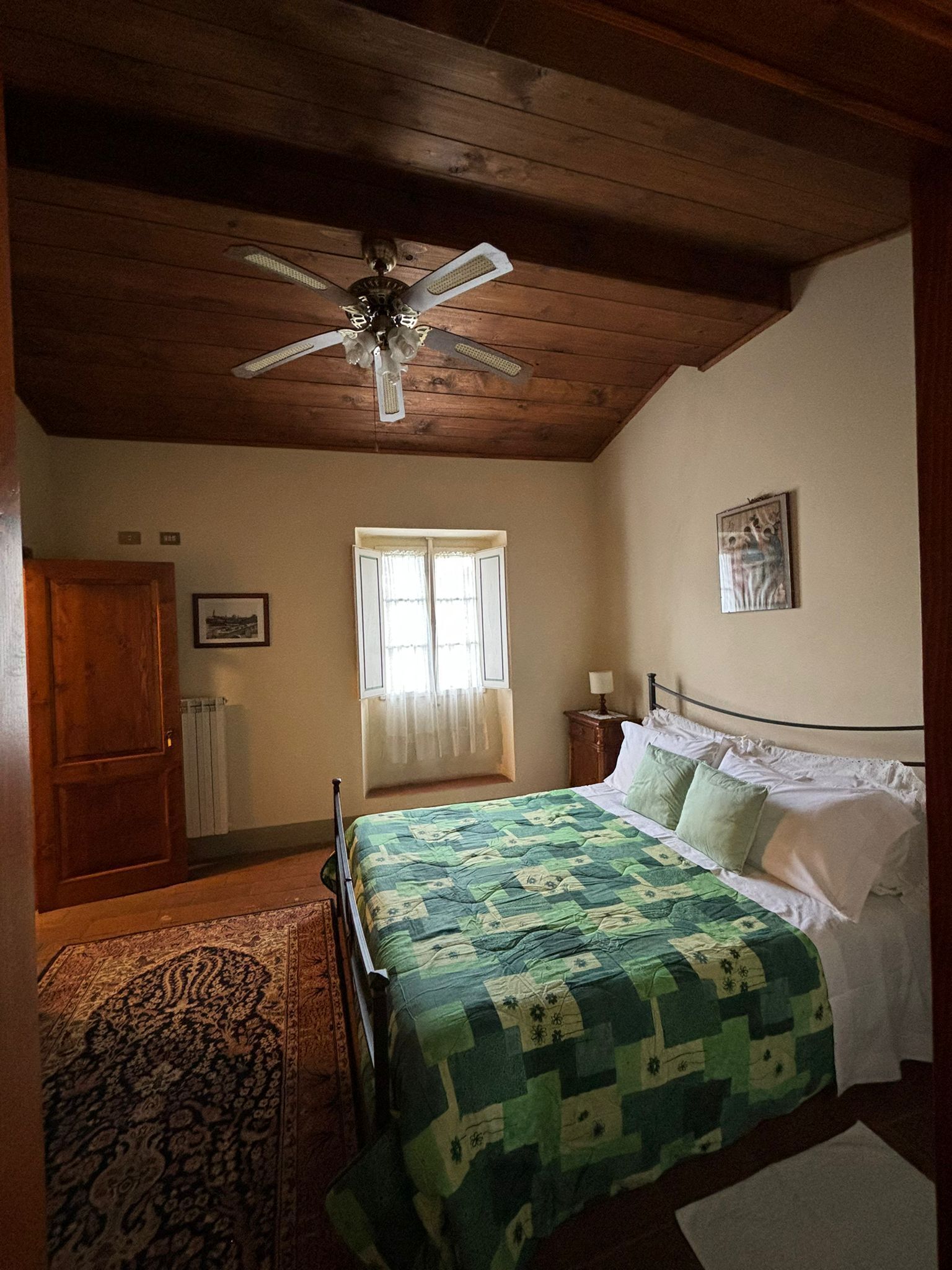 Une chambre avec un plafond en bois, un lit recouvert d’une couette verte, une table de chevet, une fenêtre avec des rideaux et un radiateur.