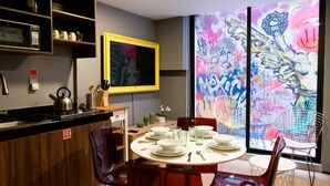Un comedor moderno con una mesa redonda preparada para cuatro personas, rodeada de sillas rojas. Un cuadro con marco amarillo adorna la pared, y se puede ver un cuadro abstracto colorido a través de un ventanal grande.
