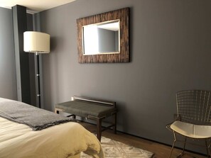 A modern bedroom with a bed, a bench, a chair, a floor lamp, and a mirror on the wall.