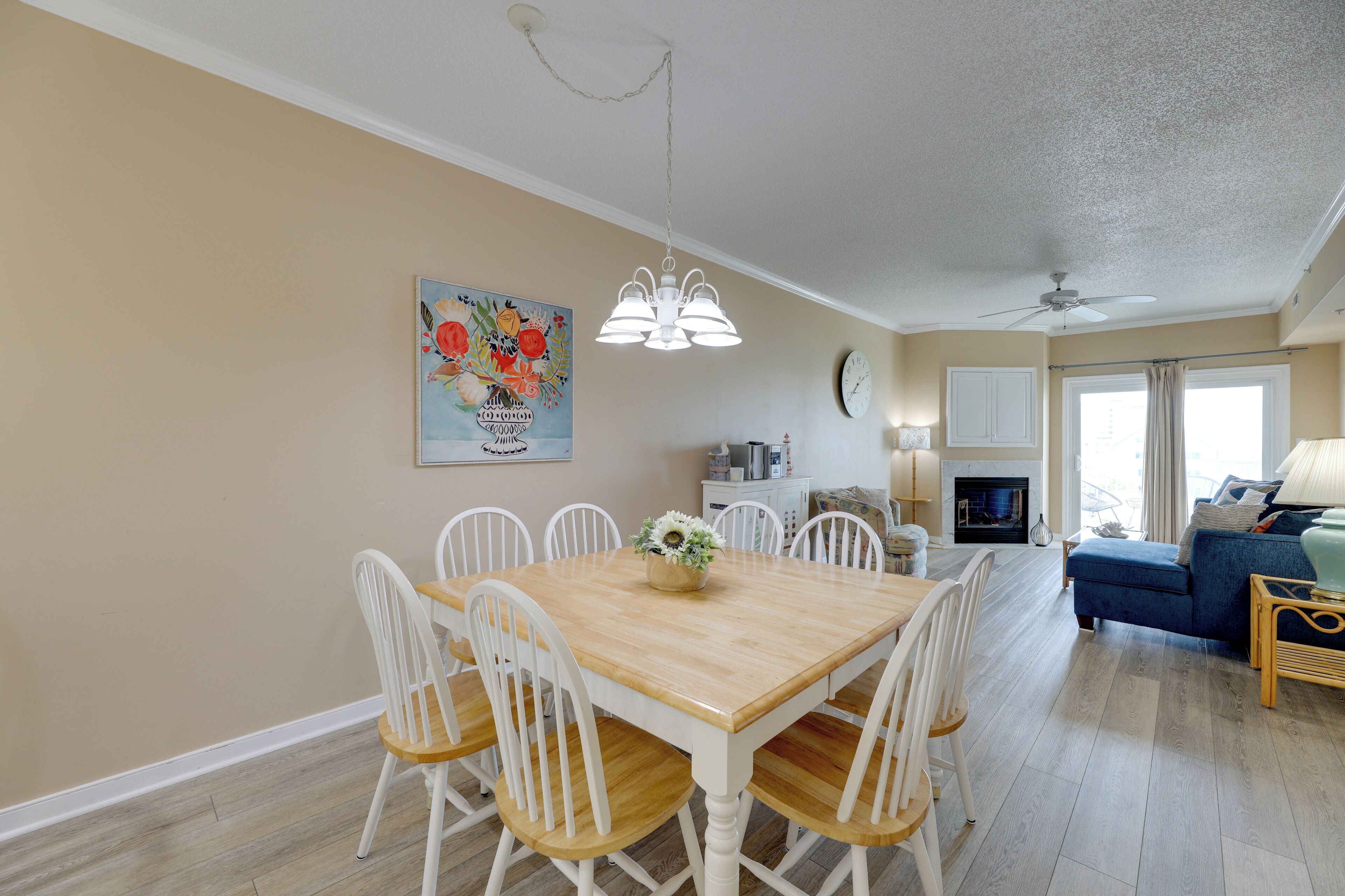 A dining area with a wooden table and white chairs, a ceiling fan, a painting on the wall, and a sofa in the background.