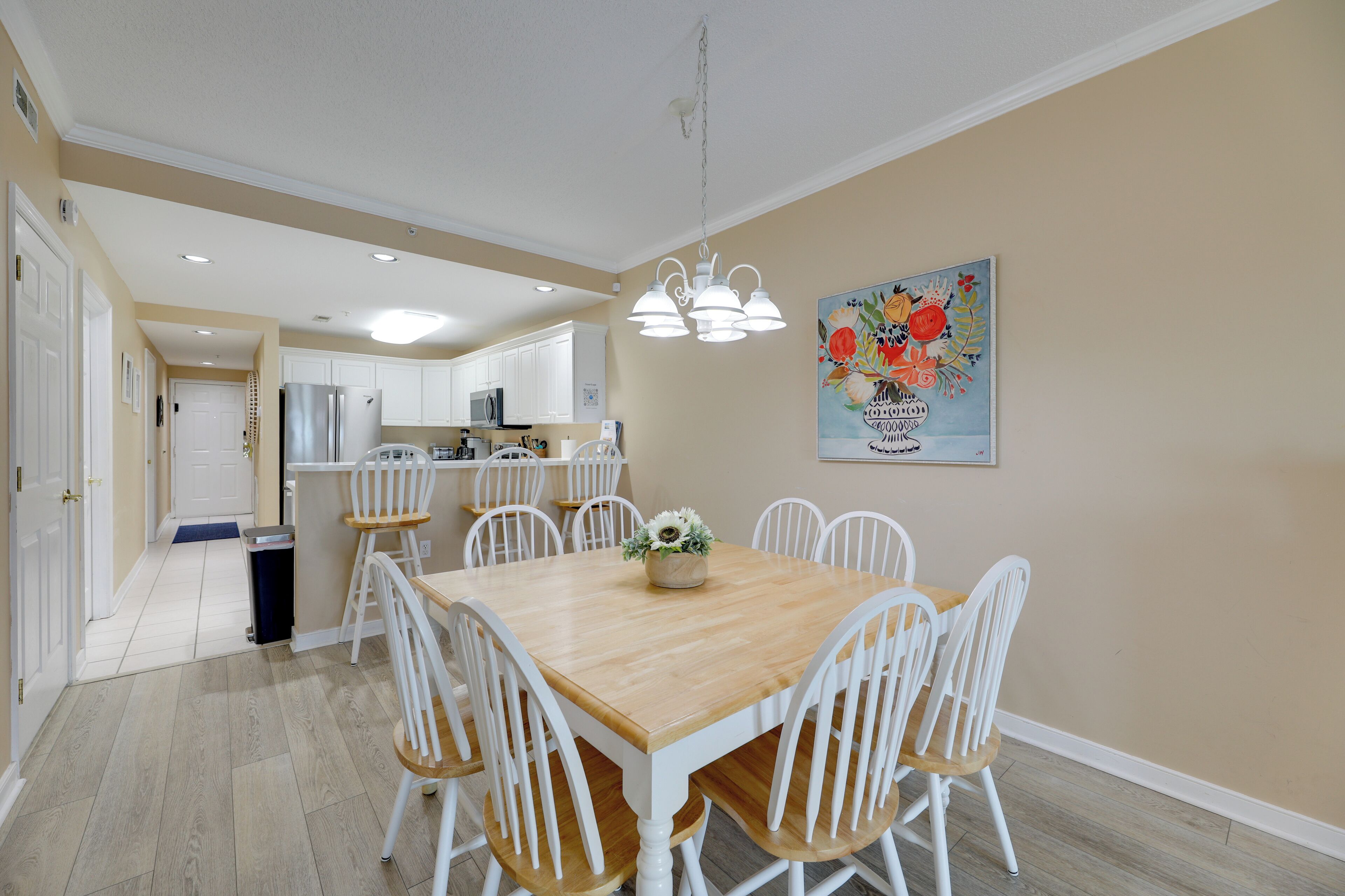 A dining area with a wooden table and white chairs, a kitchen in the background, and a painting on the wall.