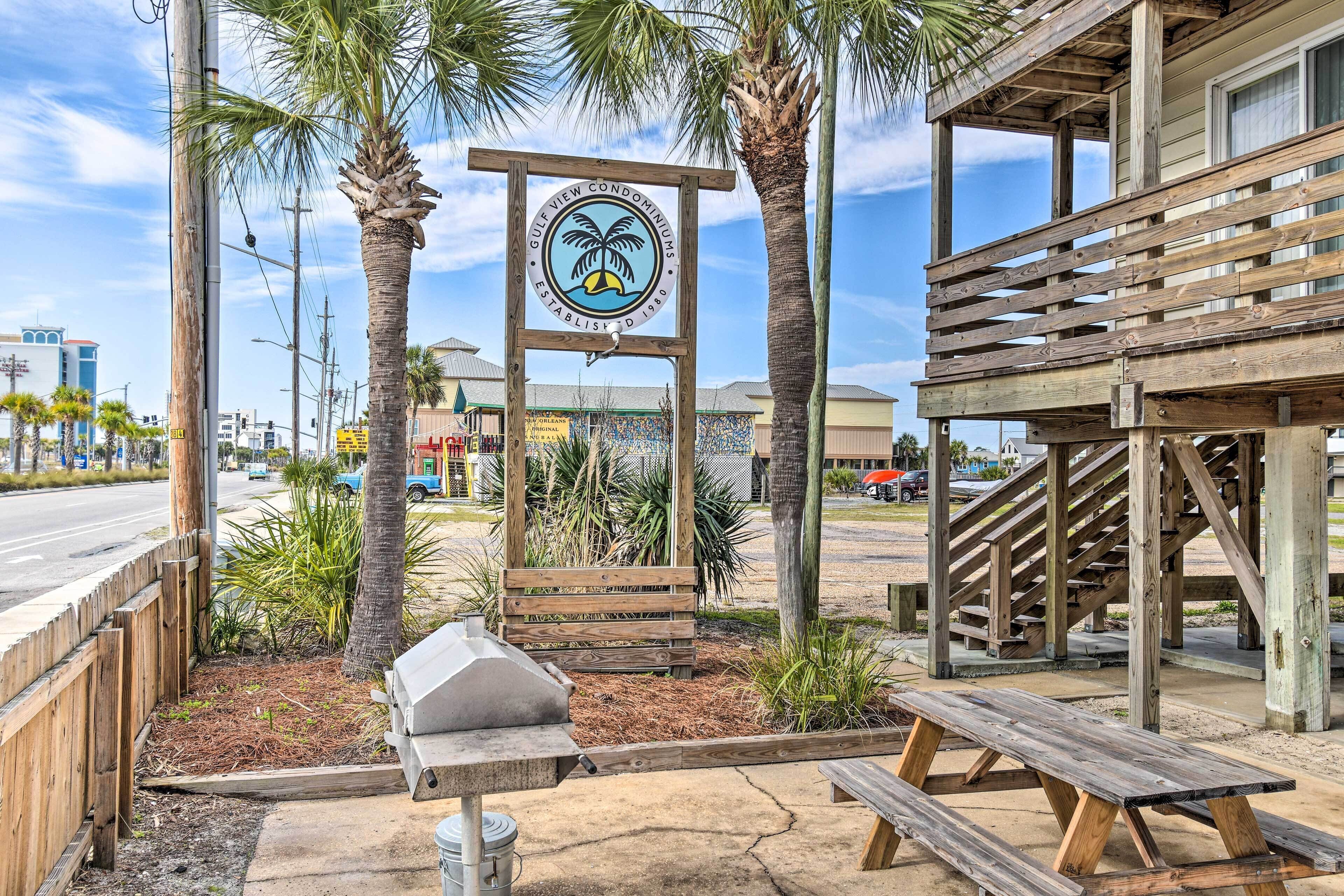A wooden sign with a palm tree logo, a barbecue grill, and picnic tables in a landscaped area with palm trees.