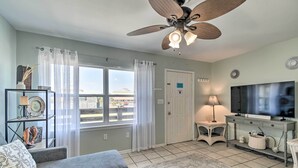 A living room with a grey sofa, a ceiling fan, a TV, and a window with curtains. - Gulf Shores Condo w/ Pool & Grill: Walk to Beach! (Gulf Shores)