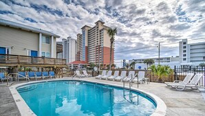 A swimming pool with lounge chairs and a wooden deck, surrounded by tall buildings. - Gulf Shores Condo w/ Pool & Grill: Walk to Beach! (Gulf Shores)