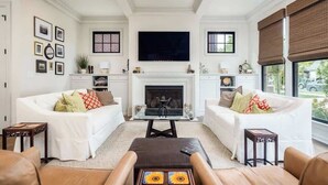 A living room with two white sofas, a fireplace, a TV mounted above it, and a large window with blinds.