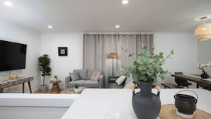A modern living room with a large TV, a grey sofa, a coffee table, and a dining area with a table and chairs. - Newly Renovated Cozy Rustic Home l 10 min from Truist Park (Smyrna)