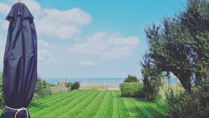 Une pelouse bien entretenue, avec vue sur la mer, une table en bois avec des chaises et un parasol bleu.