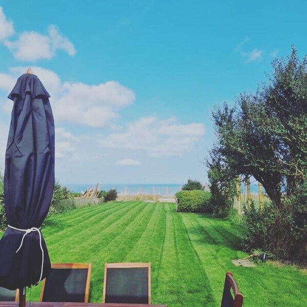 Un césped bien cuidado con vista al mar, una mesa de madera con sillas y un paraguas azul.