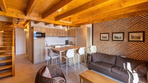 A modern kitchen with a central island, white chairs, and a brown sofa in a living area with wooden beams and herringbone-patterned wall. - Le Caribou - Appt 3 chambres proche pistes (Les Angles)
