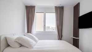 A neatly made bed with white linens and pillows, a window with curtains, and a flat-screen TV mounted on the wall.