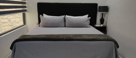 A neatly made bed with a dark headboard, white pillows, and a grey bedspread. There is a bedside table with a lamp and a small shelf. A window with blinds is visible in the background.