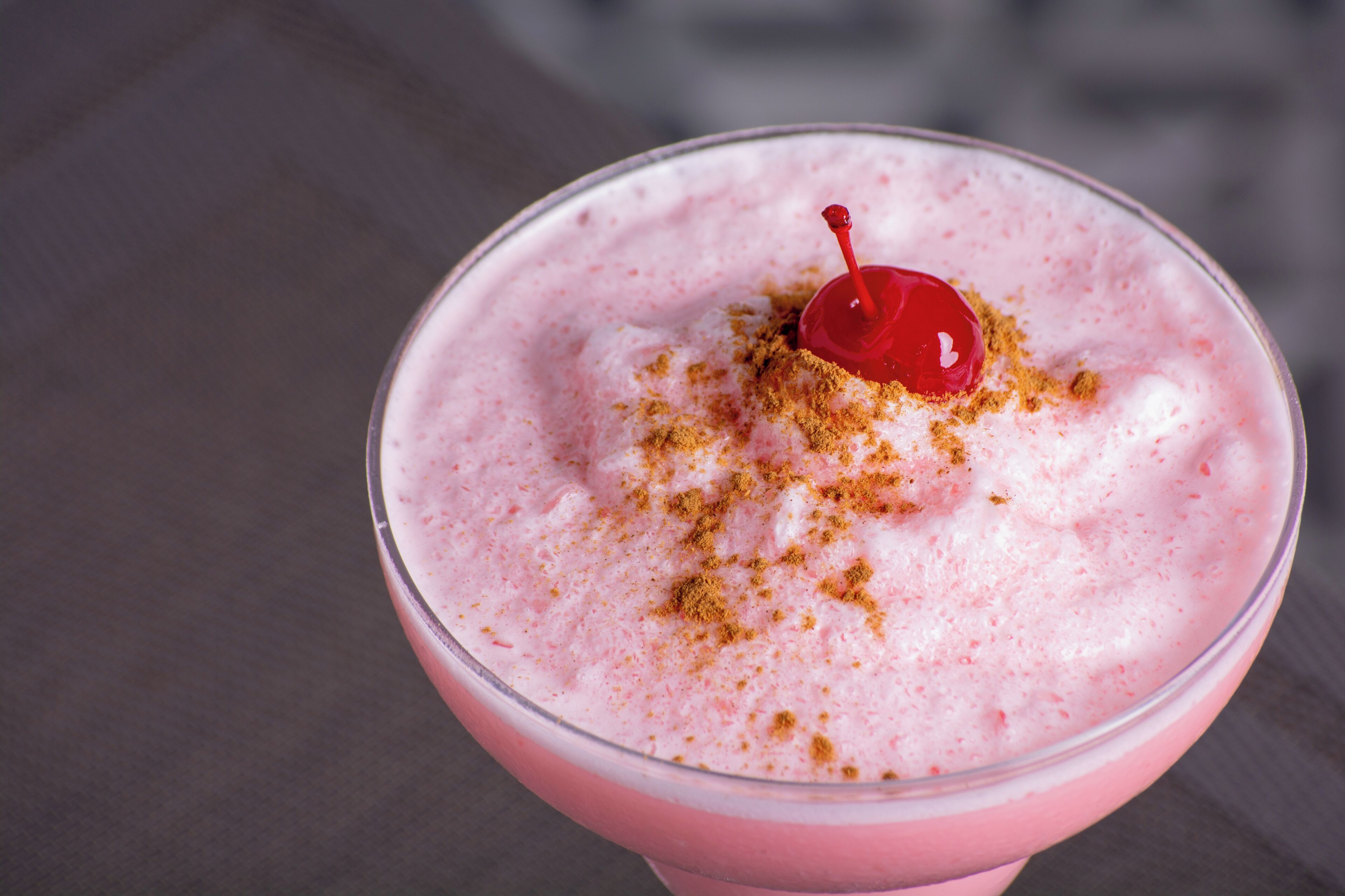 A pink beverage with a cherry and a sprinkle of a spice, served in a clear glass.