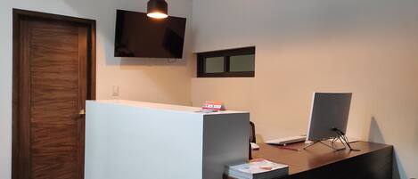 A reception desk with a flat-screen TV mounted above it, a wooden door to the left, and a small window on the wall.