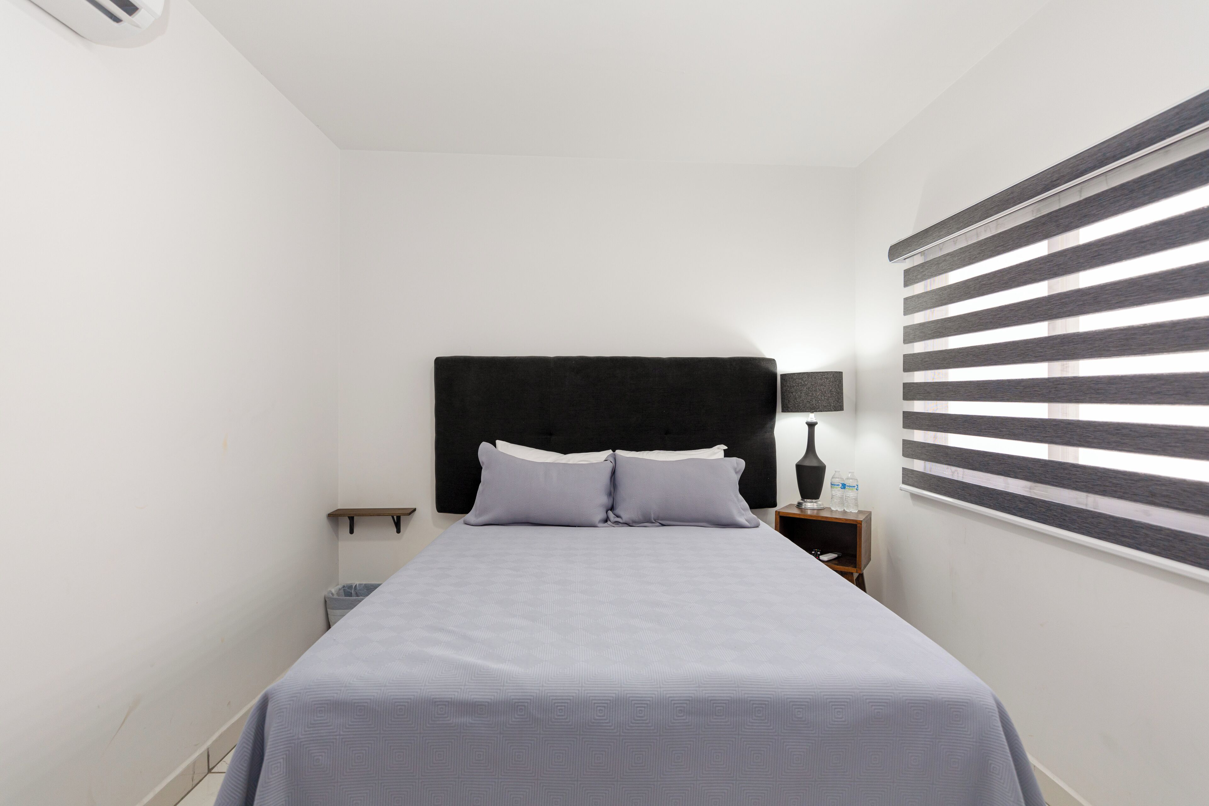 A bedroom with a bed, a nightstand, a lamp, and a window with blinds.