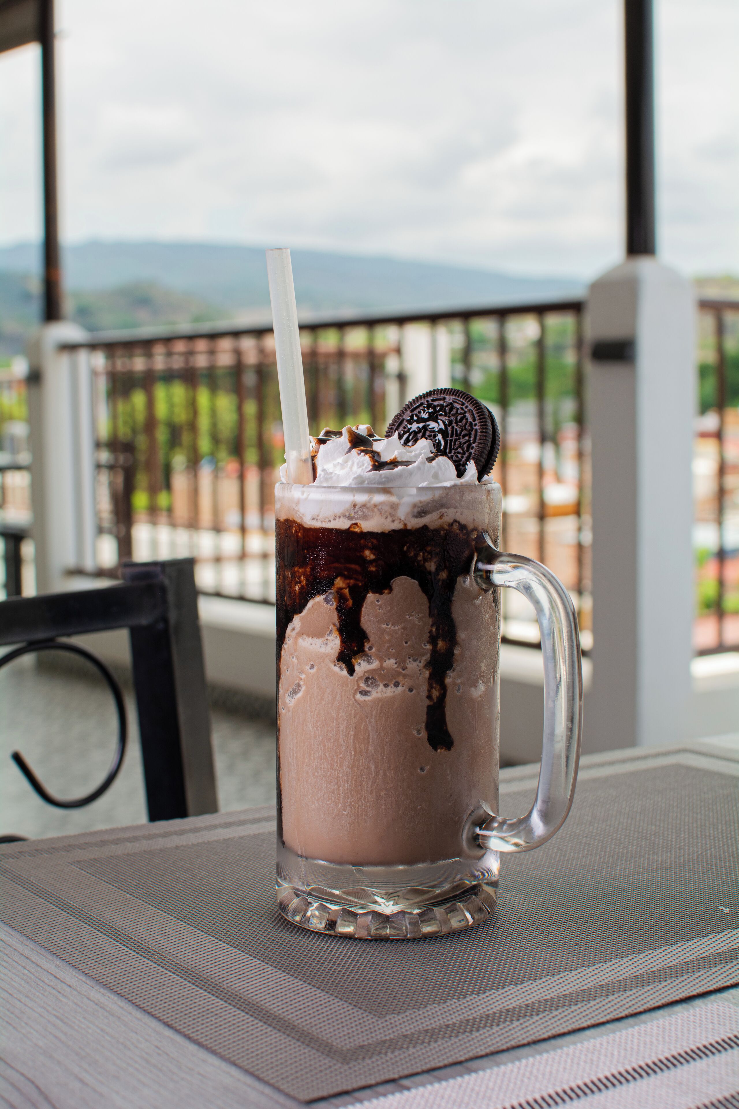 A glass mug with a straw, filled with a layered beverage, topped with whipped cream and an Oreo cookie.