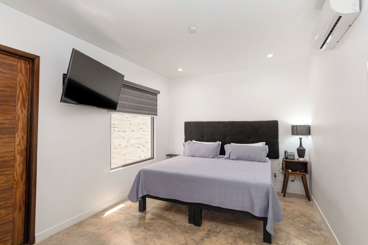 A modern bedroom with a bed, a flat-screen TV mounted on the wall, a bedside table with a lamp, and a window with blinds.