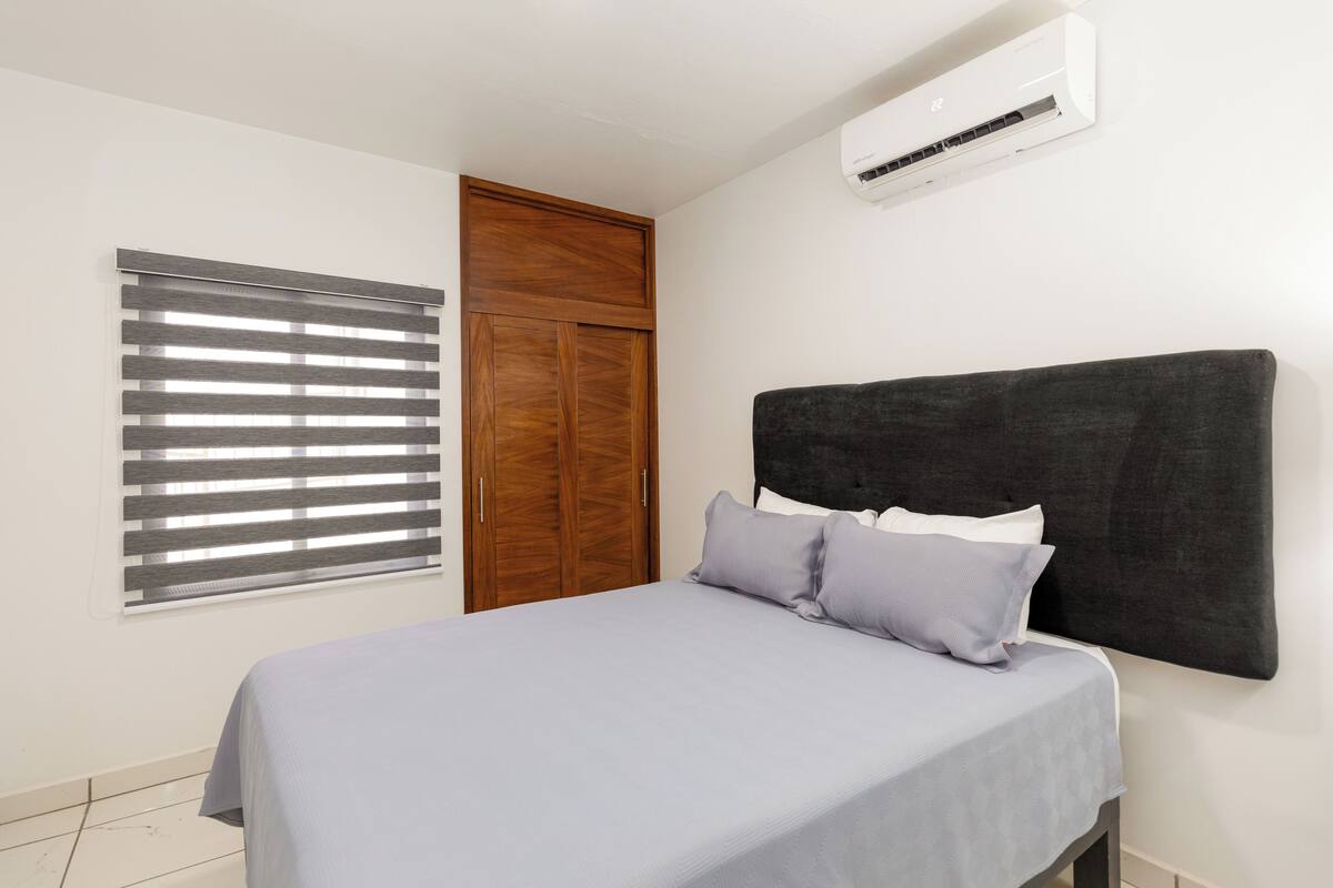 A bedroom with a bed, a wooden wardrobe, a window with blinds, and an air conditioning unit.