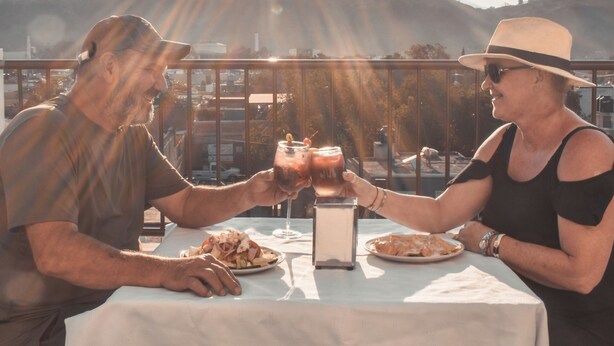 Um casal brindando com bebidas à mesa, com toalha branca, ao ar livre, com vista para colinas e céu claro.