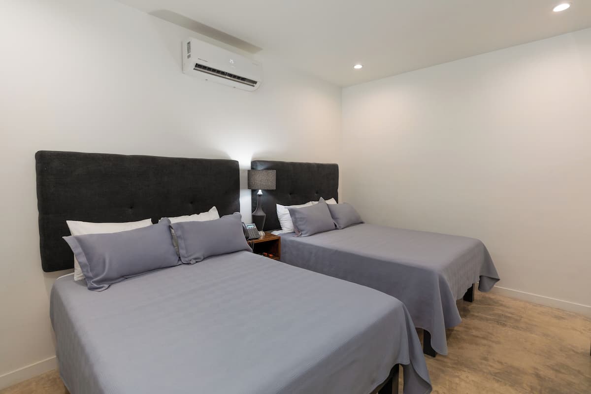 A hotel room with two beds, a nightstand with a lamp, and an air conditioning unit on the wall.