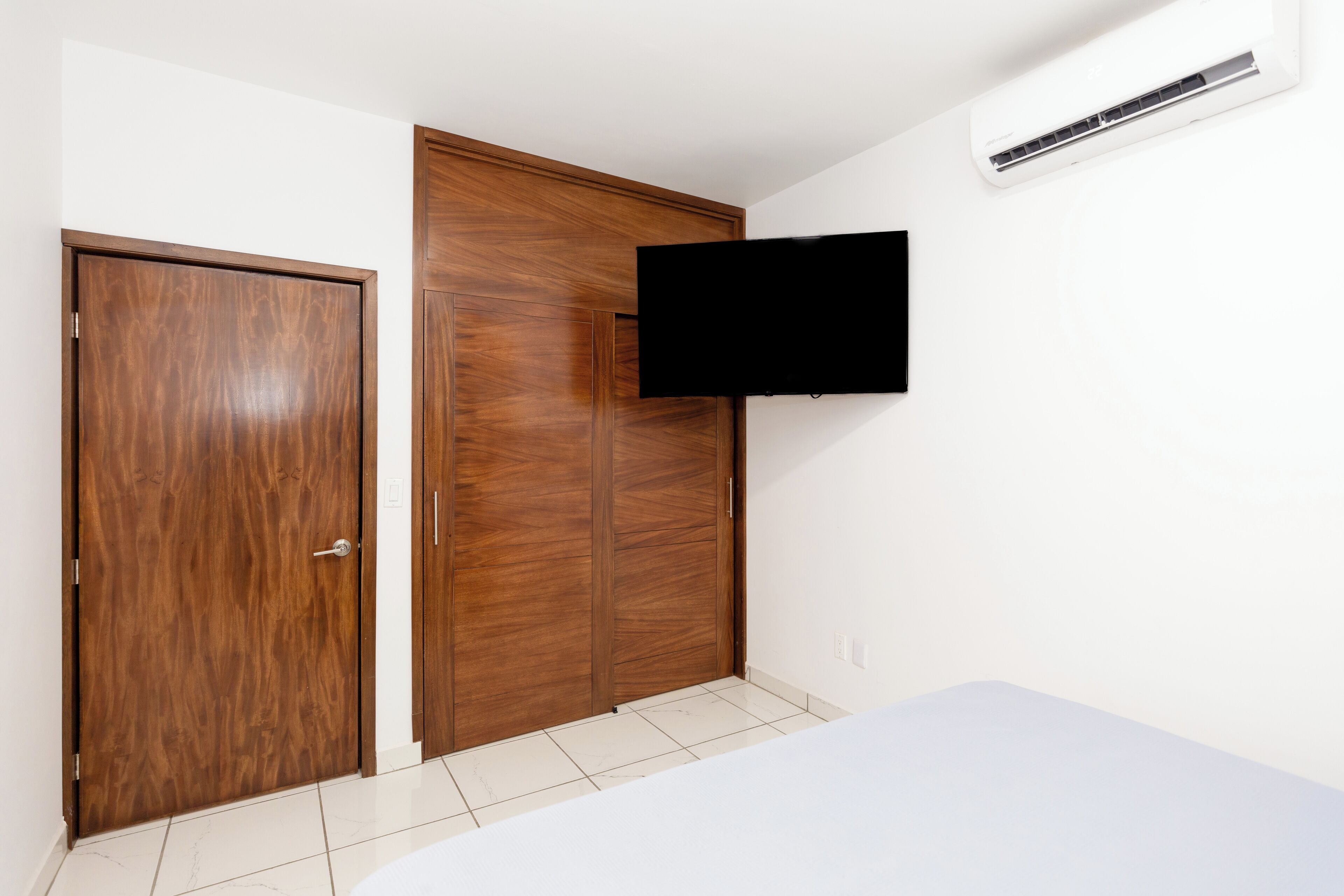Ein Schlafzimmer mit einem Holzkleiderschrank, einem Wand-Fernseher und einem weißen Bett.