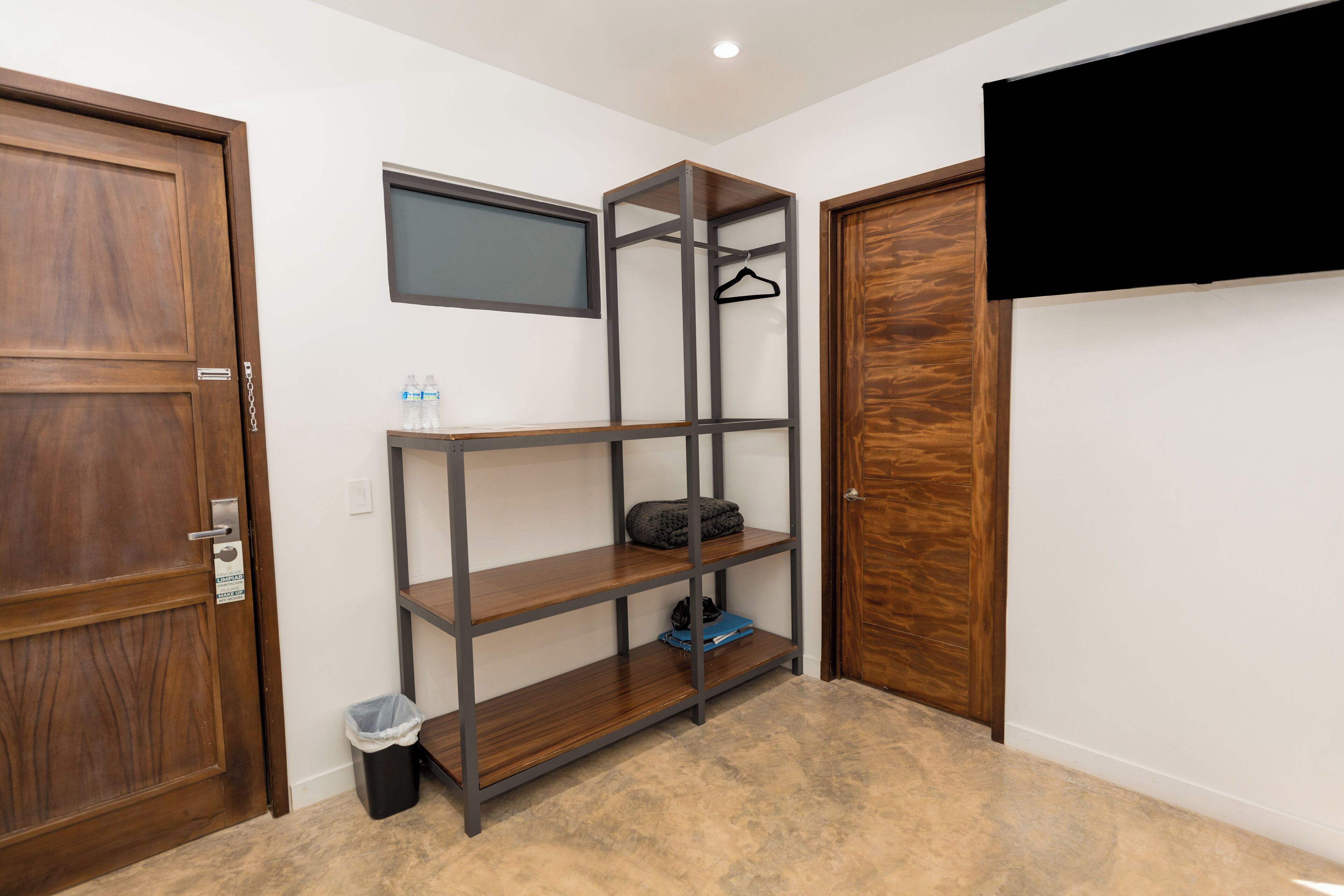 A room with a wooden door, a metal shelving unit, a small trash bin, and a television mounted on the wall.