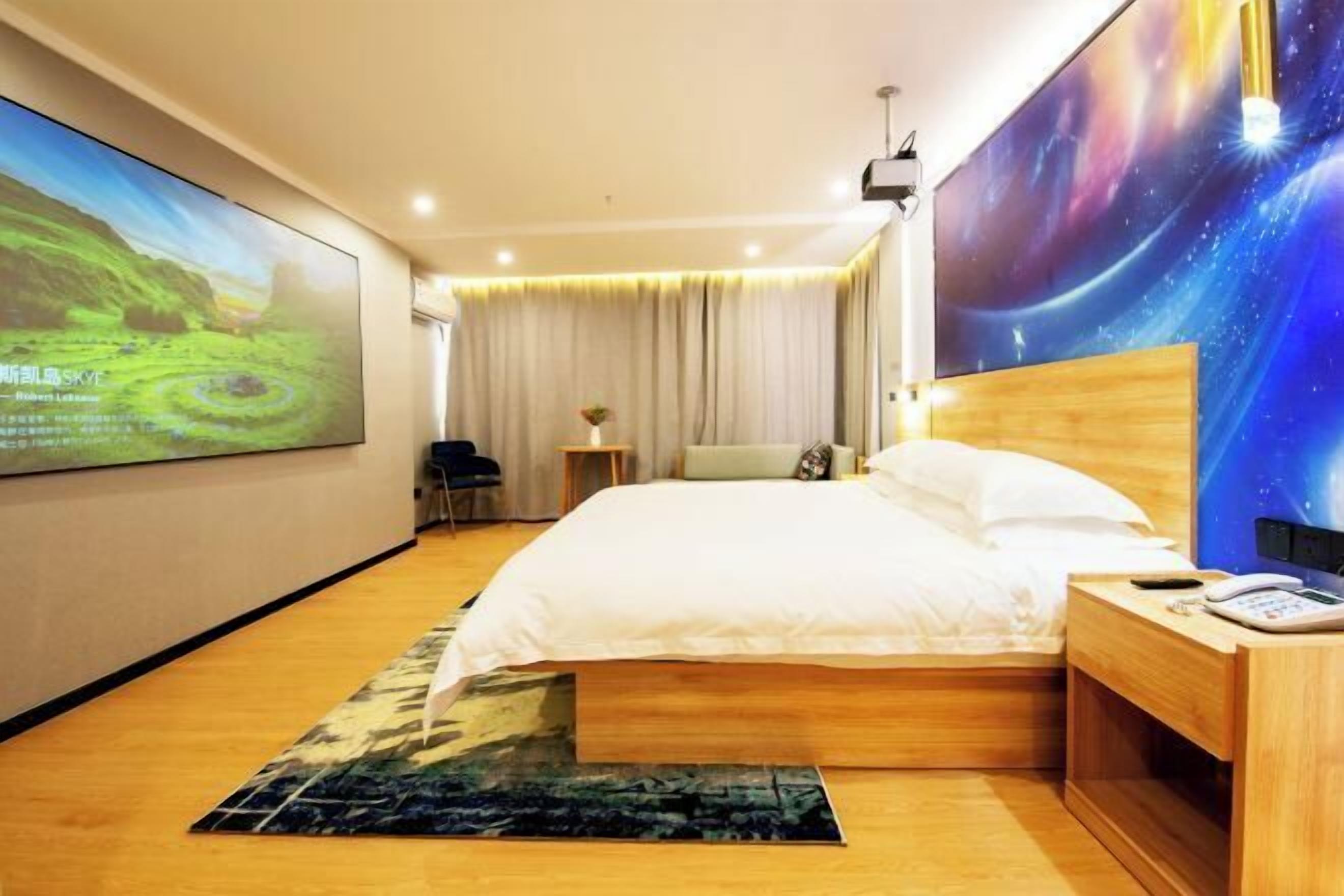 A hotel room with a large bed, a wooden headboard, a bedside table with a phone, and a wall-mounted projector screen displaying a nature scene.