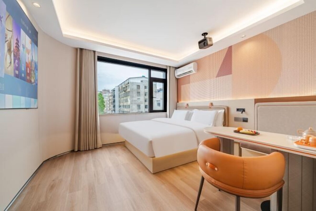 A hotel room with a bed, a desk, a chair, a window with a view of buildings, and a ceiling light.