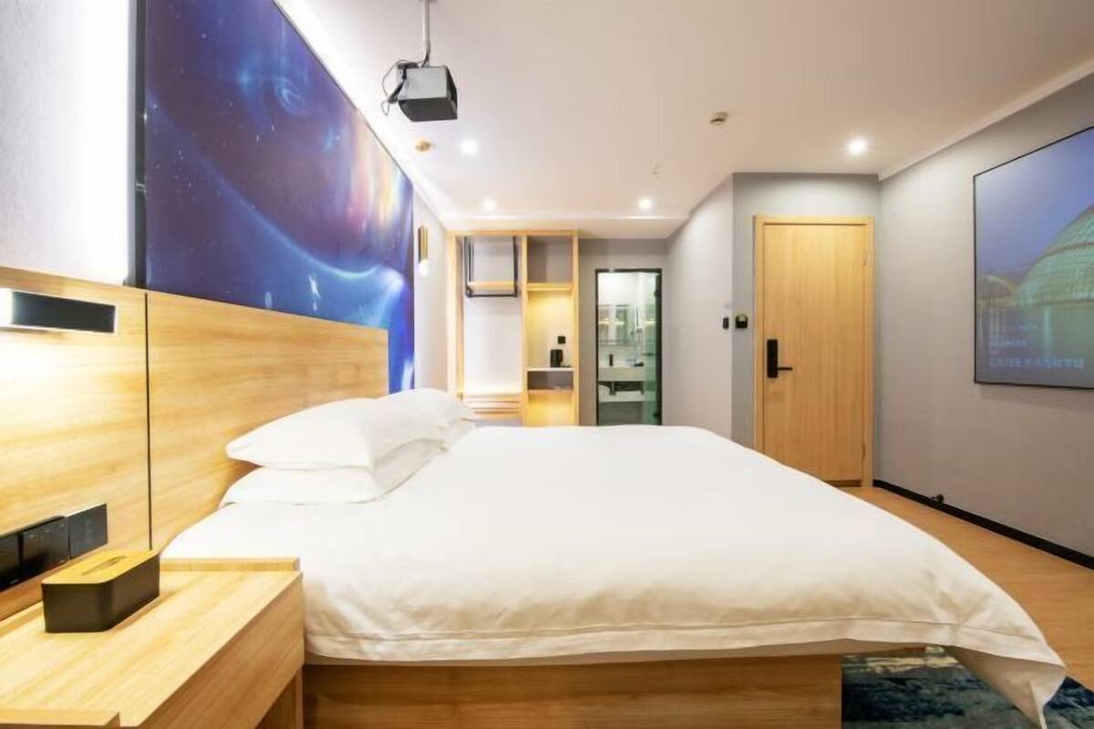 Une chambre d’hôtel moderne dotée d’un grand lit, d’une tête de lit en bois, d’une table de chevet, d’un projecteur et d’une grande fresque murale.