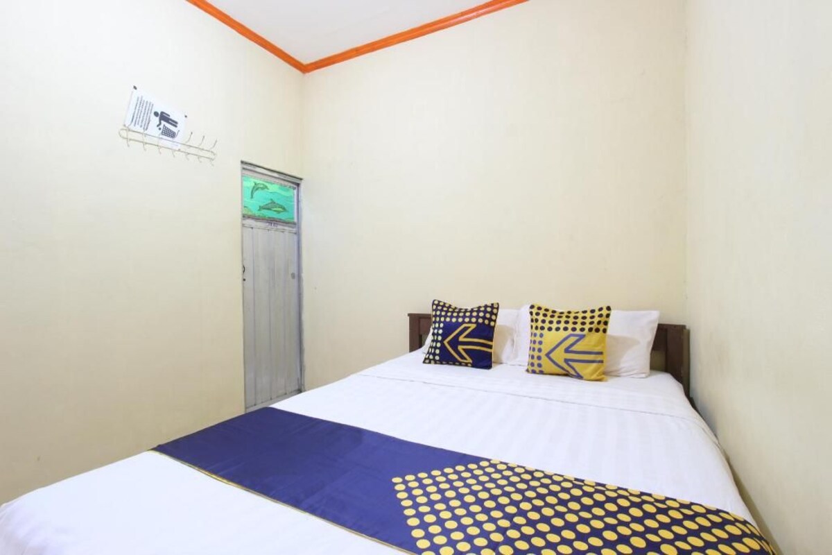 Een eenpersoonsbed met een deken met blauw en geel patroon, twee kussens met geometrische designs, en een deur met een groen licht.