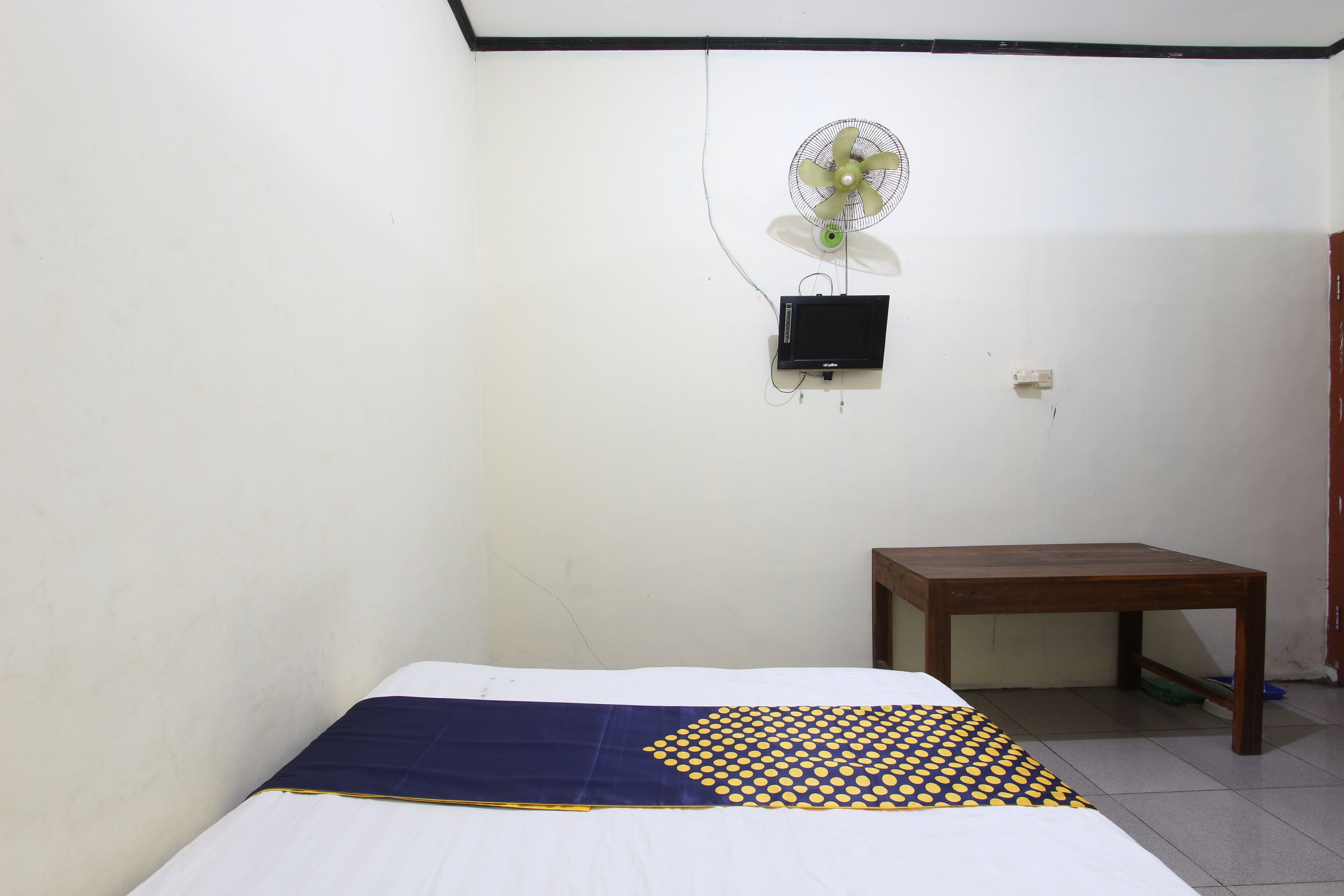 A simple room with a bed, a small wooden table, a wall-mounted fan, and a TV.